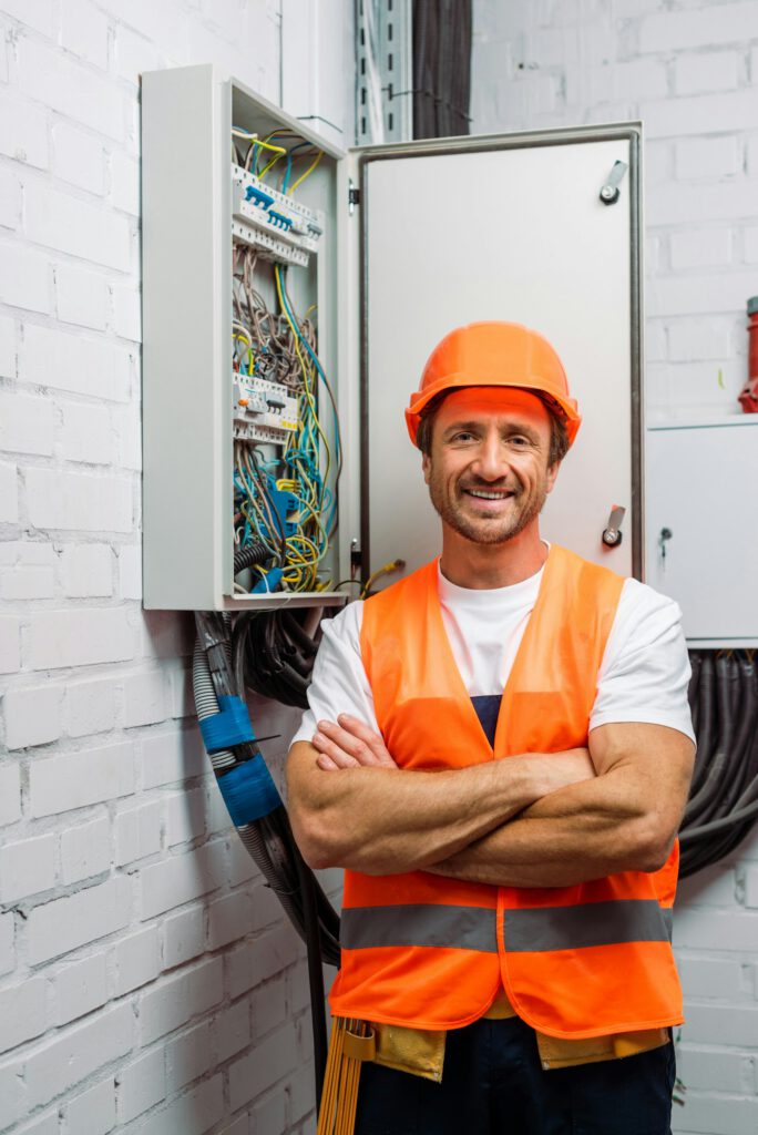 Handsome electrician in hardhat and safety vest smiling at camera near electrical distribution box